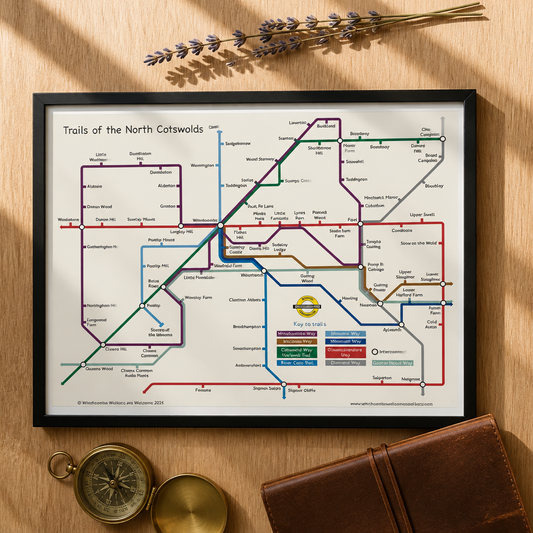 A transit map print titled 'Trails of the North Cotswolds displayed on a wooden tabletop with a vintage brass compass, a brown leather notebook, and dried lavender sprigs.